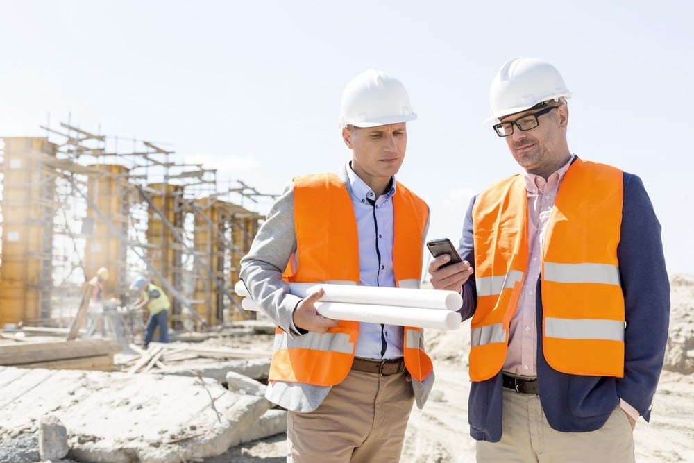 construction-contractors-3108845498 Two men in orange vests and hard hats holding papers.